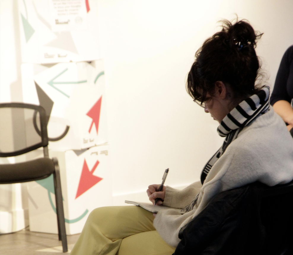 Side shot of a young lady sitting down and taking notes. Behind her is a white wall, black chair, and stack of white boxes with black, green, and red arrows (the IAF Reimagine programme branding).